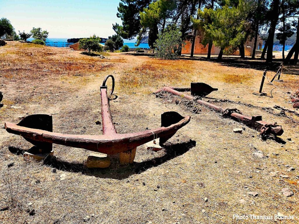 Two old rusty anchors displayed on dry ground near the sea with trees and coastline in the background