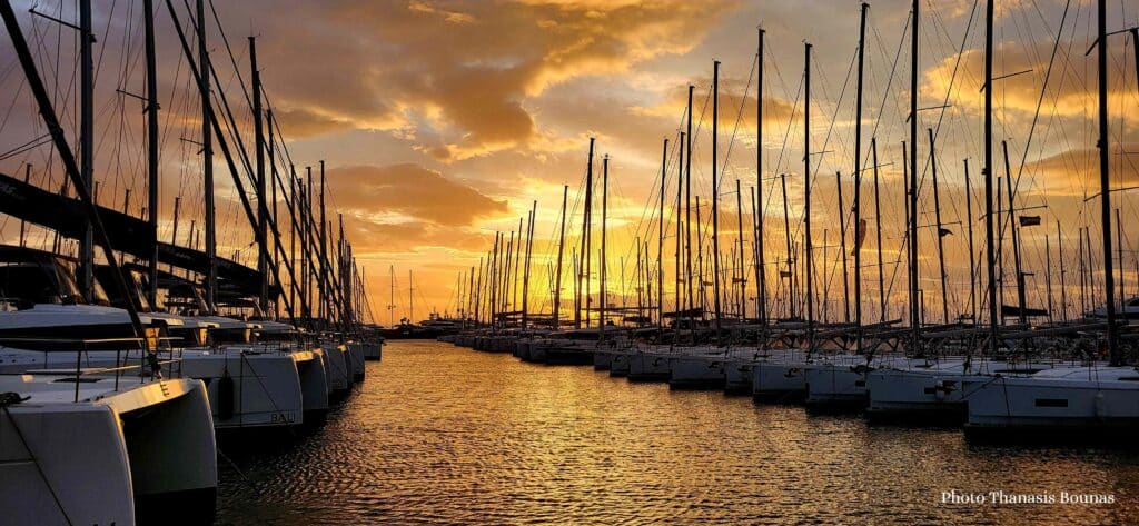 Rows of sailboats docked in a marina with tall masts silhouetted against a dramatic sunset sky