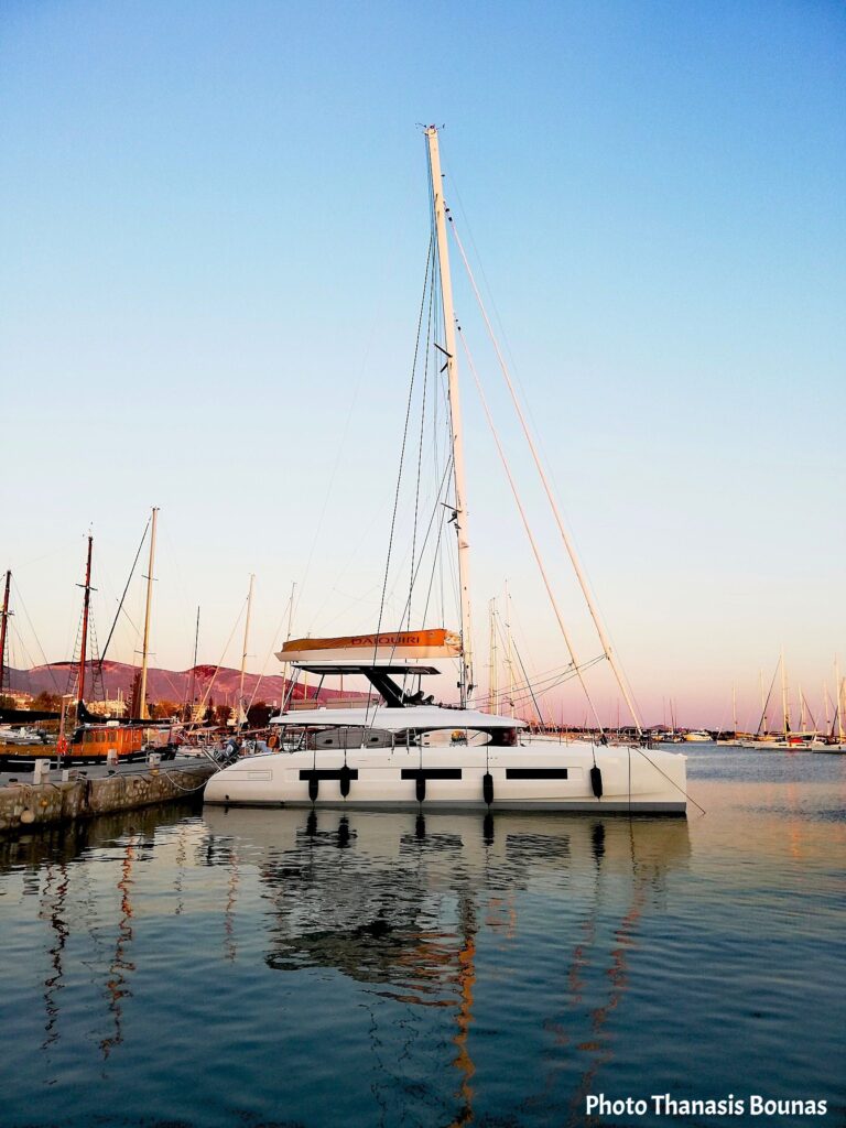 White catamaran sailboat docked in a marina during sunset with calm water reflecting the boat and tall mast